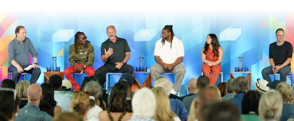 A wide shot of a panel discussion at the Aspen Ideas Festival. Six people are seated on stools on a stage against a bright blue geometric backdrop featuring the festival's logo. Chris Varelas, Cornelius Wills, Tim Pearce, Tyrone Riley Sr., Emily Avalos, and Michael Soenen are engaged in conversation, with Tim Pearce gesturing as he speaks. In the foreground, the blurred backs of a large audience are visible, watching the presentation.